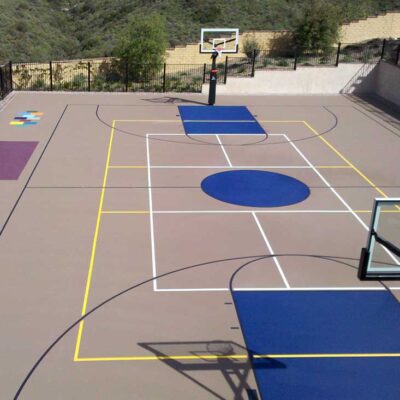 The Photo Shows A High Elevation View Of A Shared Multi-sport Court In Beige With Black Lines For Basketball (plus Solid Blue Key And Center Circle), Yellow Volleyball Lines, And White Pickleball Lines.