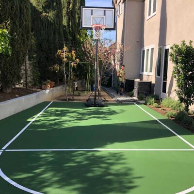 The Photo Shows Two Large Buildings With Just Enough Room To Fit A Basketball Court In Green With White Striping In Between Them.