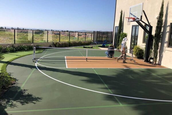 The Photo Shows A Half Basketball Court On The Side Of A Big Residence With Medium Green Court And Surround, Beige In The Court's Key Area And White Striping.
