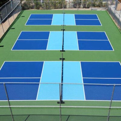 Photo Shows Three Pickleball Courts In Blue With Light Blue Kitchens And Green Surrounds.