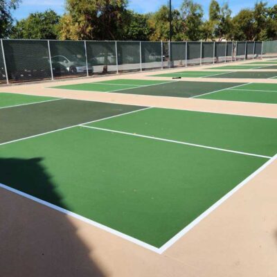 the photos shows six pickleball courts in a row with green courts, dark green kitchens, white striping and a sandstone colored surround.