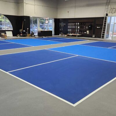 the photo shows two indoor pickleball courts constructed by ferandell tennis courts using a combination of blue and light blue for the courts and gray for the surround.