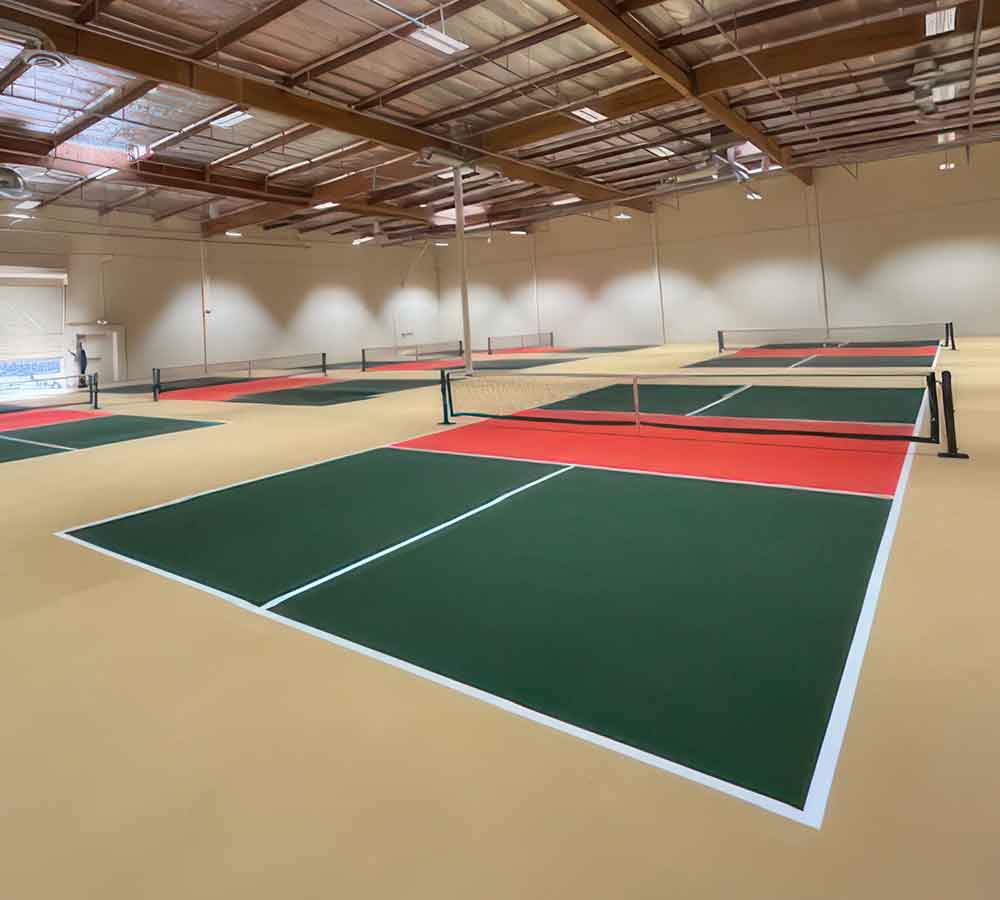 photo shows multiple green & orange indoor pickleball court with tan surround.